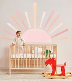 a baby standing in a crib next to a rocking horse and sun mural on the wall