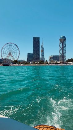 a view from the back of a boat in the water with buildings and ferris wheel in the background