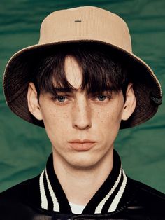 a young man with freckled hair wearing a brown hat and black shirt against a green background