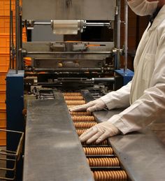 a man in white jacket and mask standing next to conveyor belt with hotdogs on it