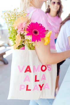 a woman carrying a shopping bag with flowers in it and the words vamos al la playa printed on it