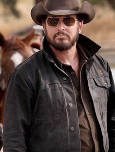 a man wearing a cowboy hat and leather jacket standing in front of a brown cow