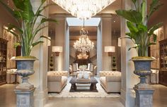 a living room filled with lots of furniture next to tall potted plants and chandeliers