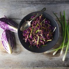 a bowl filled with red cabbage and green onions next to two spoons on top of a wooden table