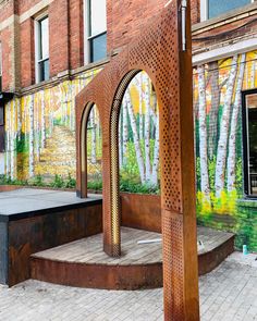 a large metal structure sitting on top of a brick floor next to a building with trees painted on it