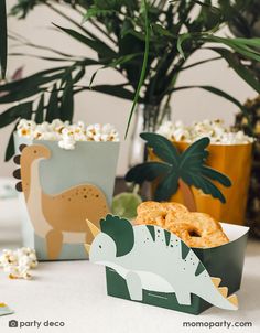 two dinosaur boxes filled with doughnuts and sprinkles next to potted plants