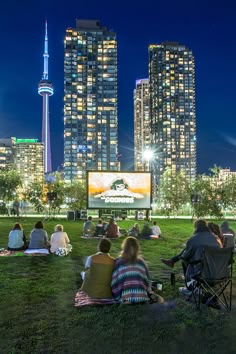 people are sitting in the grass watching movies at night with tall buildings in the background