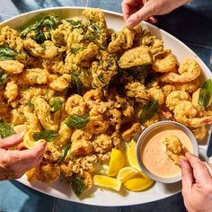 two hands reaching for some fried food on a white plate with lemon wedges and ketchup