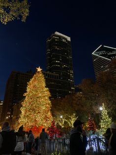 the christmas tree is lit up at night with people standing around it and buildings in the background