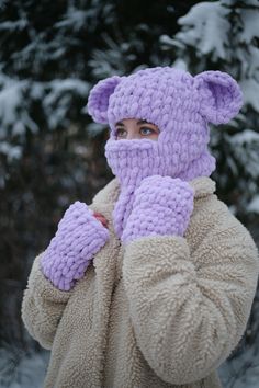 a woman wearing a purple knitted bear hat and mittens while standing in the snow