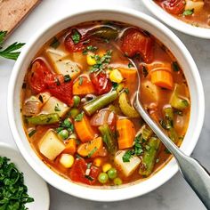 two bowls filled with vegetable soup on top of a white table next to bread and vegetables