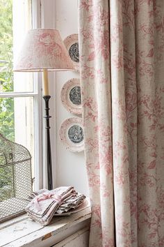 a window with pink curtains and plates on the windowsill, next to a lamp