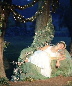 a woman in a white dress is laying on a green chair under the tree with fairy lights