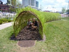 two people sitting on the ground in front of a bamboo arch with grass growing around it