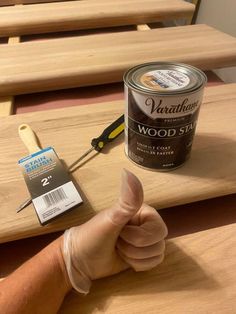 a hand holding a paintbrush next to a can of wood stain