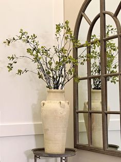 a white vase with some green plants in it sitting on a table next to a mirror