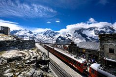 a train traveling down tracks next to snow covered mountains in the distance with people standing on top