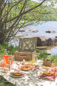 a picnic table with food and drinks on it near the water in front of some trees