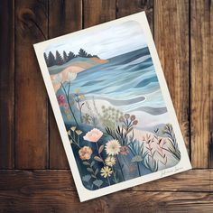 a card with an image of the ocean and flowers on it, against a wooden background