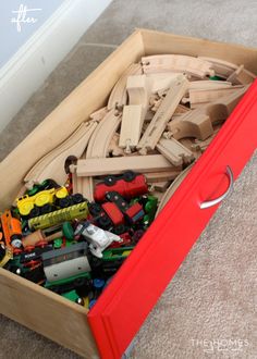 a red box filled with wooden toys on top of carpeted floor next to wall