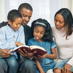 three adults and two children sitting on a couch looking at an open book while the child is reading it