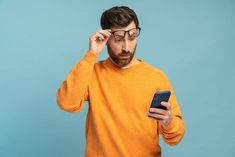 a man in an orange sweater is looking at his cell phone and holding it to his forehead