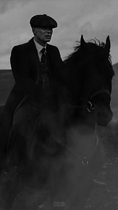 a man riding on the back of a black horse next to a field and cloudy sky