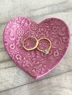 two gold wedding rings are placed on top of a pink heart - shaped box with floral designs