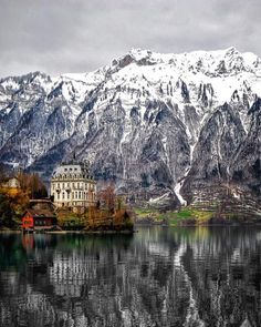 an old building on the shore of a lake with snow covered mountains in the background