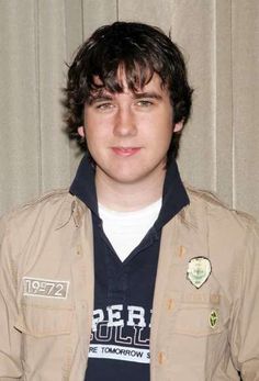 a young man wearing a brown jacket standing in front of a curtain