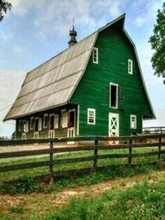 a large green barn sitting on the side of a lush green field next to a wooden fence