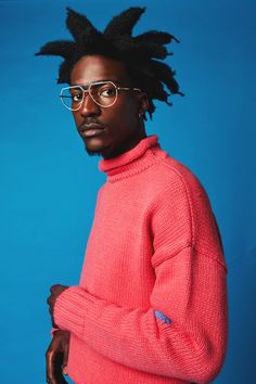 a man with dreadlocks wearing a pink sweater and glasses, standing in front of a blue background