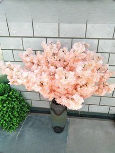 a vase with pink flowers sitting on the ground next to a brick wall and shrubbery