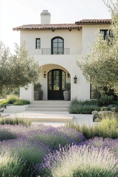 a white house with lots of lavender bushes and trees in front of the entrance to it