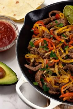 a skillet filled with meat and vegetables next to an avocado, tortilla chips and salsa