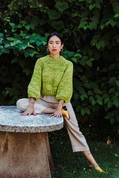 a woman sitting on top of a cement bench in front of trees and bushes wearing a green sweater