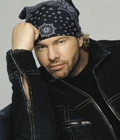 a man wearing a black jacket and bandana on top of his head, sitting in a chair