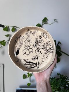 a hand holding a ceramic bowl with designs on the inside and outside, in front of a potted plant