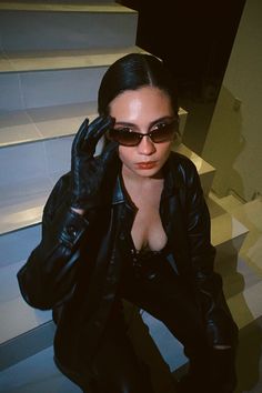 a woman in black jacket and sunglasses sitting on stairs with her hand up to her face