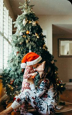 a woman sitting in front of a christmas tree kissing her baby's nose with a santa hat on