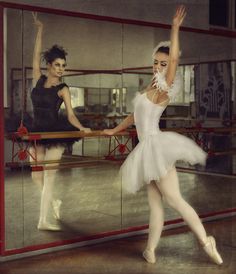 two ballerinas in white tutus and black leotards, one is holding her leg up