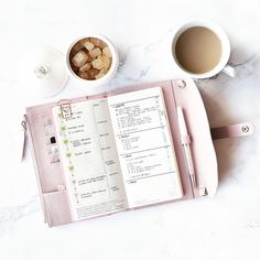 an open pink planner next to two cups of coffee and a pen on a marble table