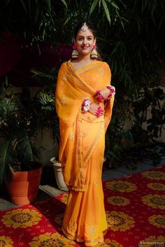 a woman in an orange sari standing on a red and yellow carpet with potted plants