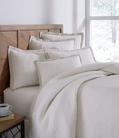 a bed with white comforters and pillows in front of a window, next to a wooden headboard