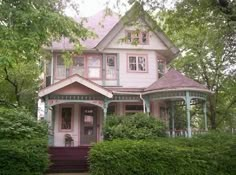 a pink house is surrounded by green bushes and trees in front of it, with a porch on the second floor