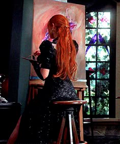 a woman with long red hair sitting in front of an easel