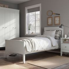 a white bed sitting in a bedroom next to a dresser and window on top of a hard wood floor