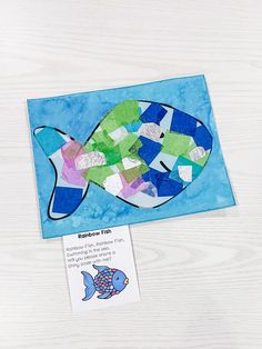 a fish made out of colored paper on top of a white wooden table next to a card