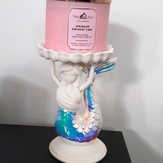 a pink candle holder with a mermaid holding a birthday cake on it's pedestal