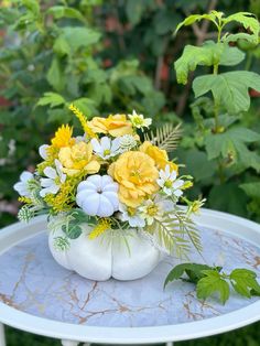 a white vase filled with yellow flowers sitting on top of a metal table next to green leaves